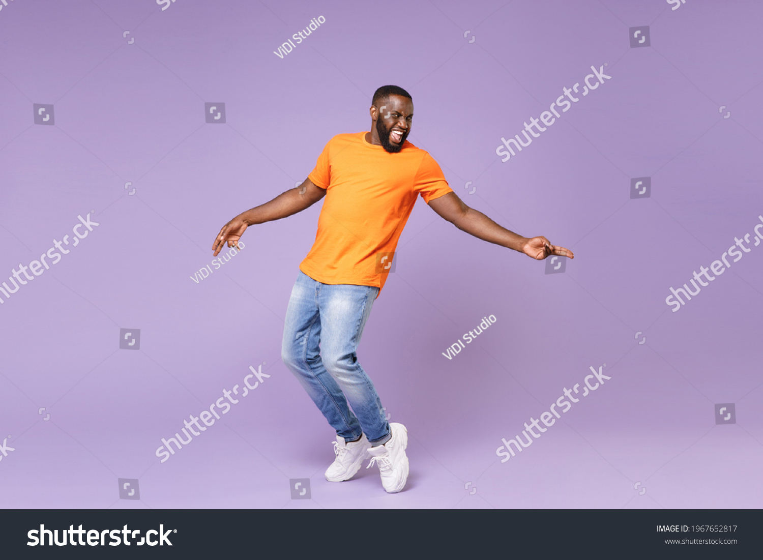 Full length of excited funny young african american man 20s in basic casual orange t-shirt dancing standing on toes pointing index fingers aside isolated on pastel violet background studio portrait Stock-foto