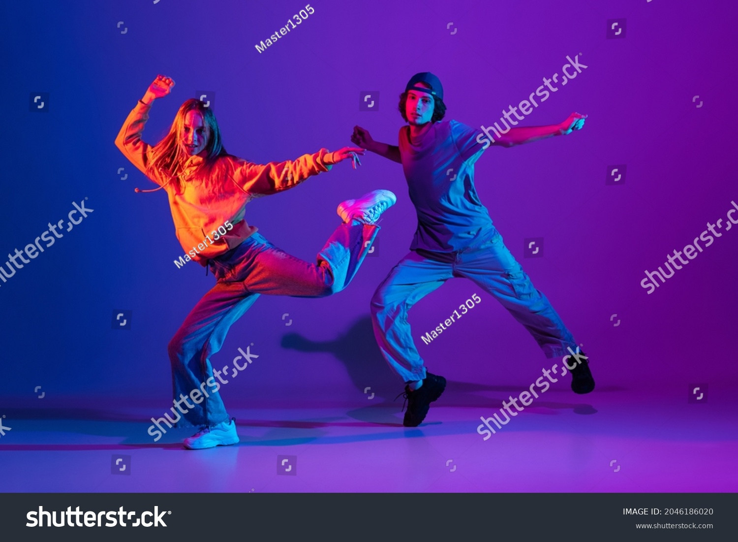 Stylish man and woman dancing hip-hop in casual sports youth clothes on gradient purple pink background at dance hall in neon light. Youth culture, hip-hop, movement, style and fashion, action. Stock-foto
